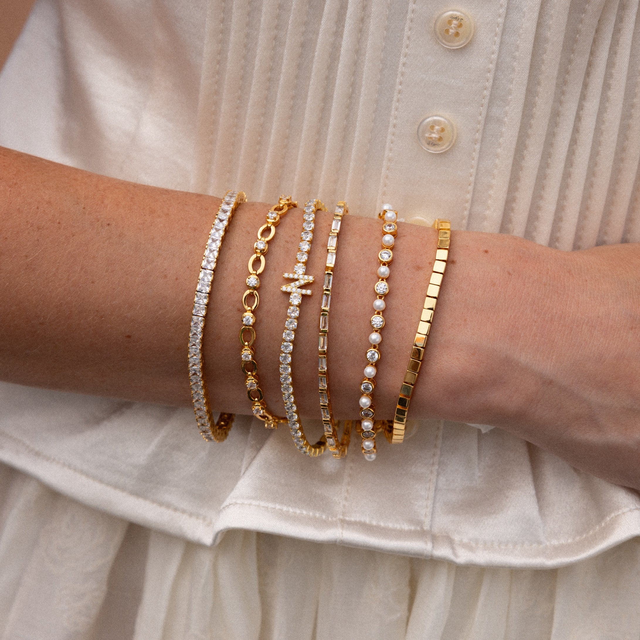 A womans arm showcases five bracelets, including the Diamond Chain Tennis Bracelet, with gold and diamond links, all set against a white buttoned top.
