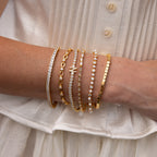 A person wears a white top and several gold and diamond bracelets, including the Princess Diamond Tennis Bracelet in 18K Gold, adorned with sparkling princess-cut diamonds.