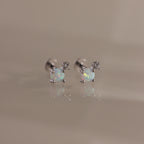 Studio shot of a pair of silver flatback stud earrings, each set with a round opal gemstone and a small diamond accent on a reflective surface.