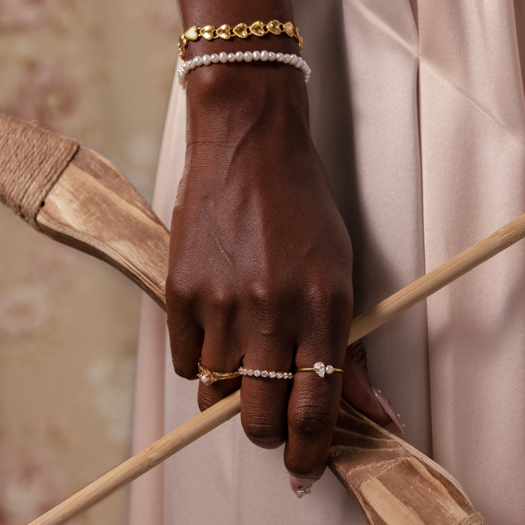 A hand wearing the Ribbed Heart Link Bracelet in 18K Gold holds two wooden arrows against a soft beige background.