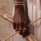 A hand wearing the Vintage Pave Pearl Ring and gold jewelry holds a wooden bow against a soft beige background.
