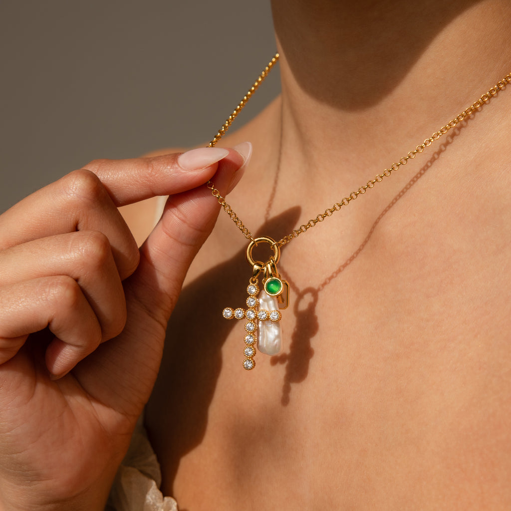 A hand holds the Long Baroque Pearl Charm in 18K Gold, a necklace featuring a jeweled cross, personalized charm, green accent, and stunning pearls worn around a womans neck.