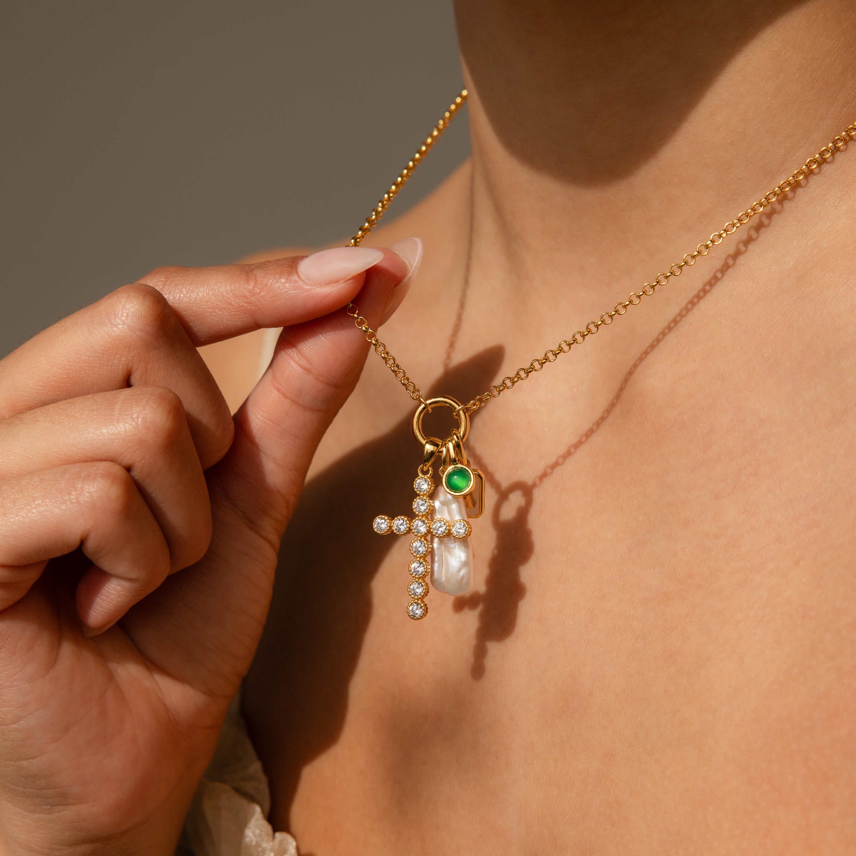 A hand holds the Long Baroque Pearl Charm in 18K Gold, a necklace featuring a jeweled cross, personalized charm, green accent, and stunning pearls worn around a womans neck.