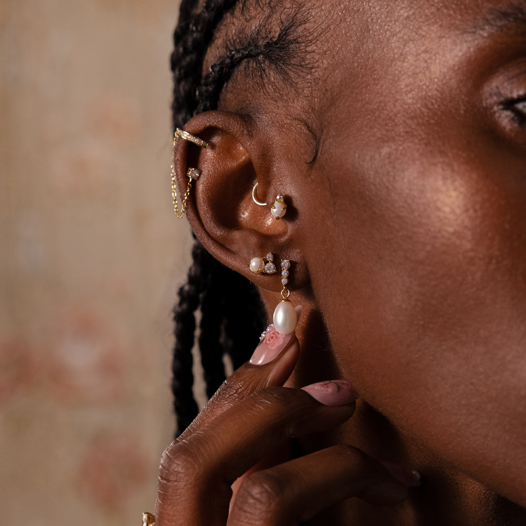 A close-up of a person with braided hair wearing multiple gold and pearl earrings, including the Millie Drop Earrings in 18K Gold, gently touching one earring.