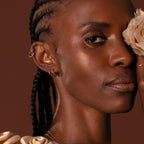 Woman with braided hair wears Inside Out Birthstone Huggies and a cream rose covering one eye, set against a brown background.
