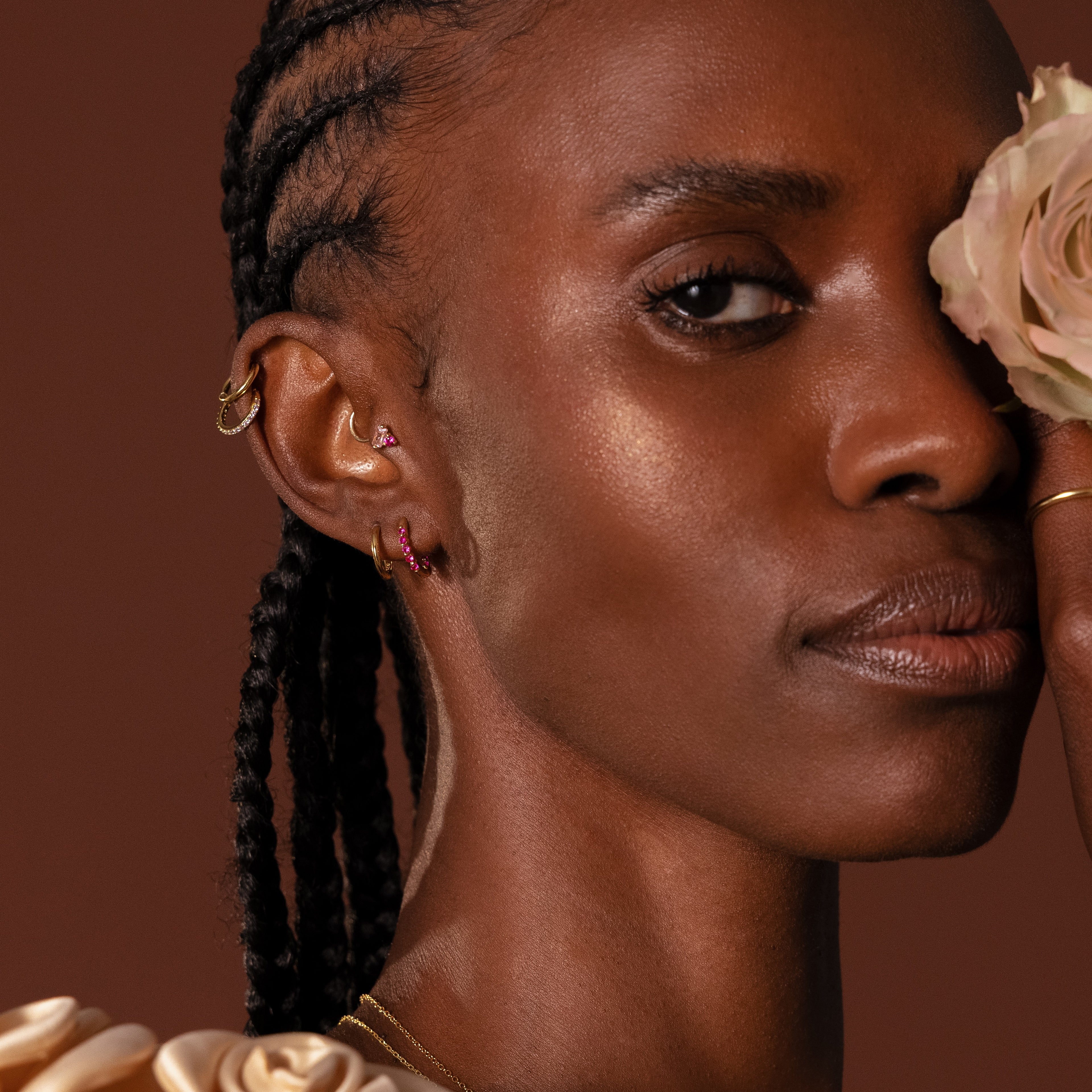 Woman with braided hair wears Inside Out Birthstone Huggies and a cream rose covering one eye, set against a brown background.