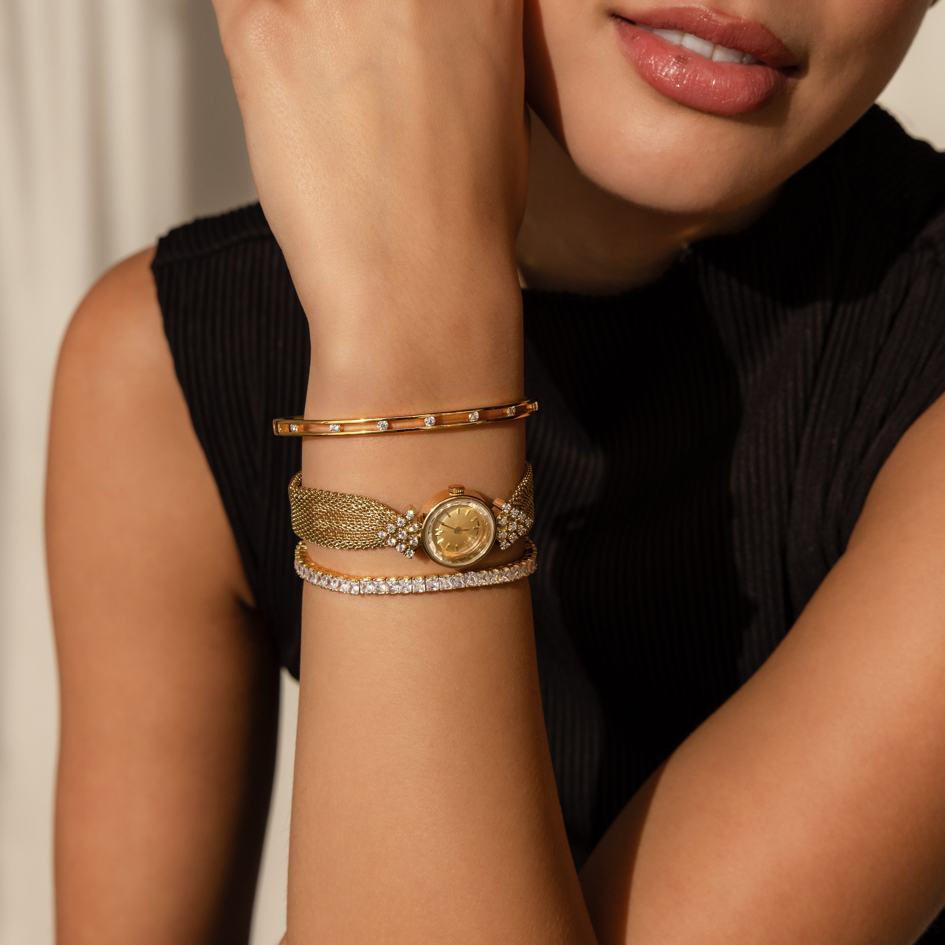 A woman in a black top showcases the Diamond Mesh Timepiece and gold bracelets, her hand thoughtfully near her chin.