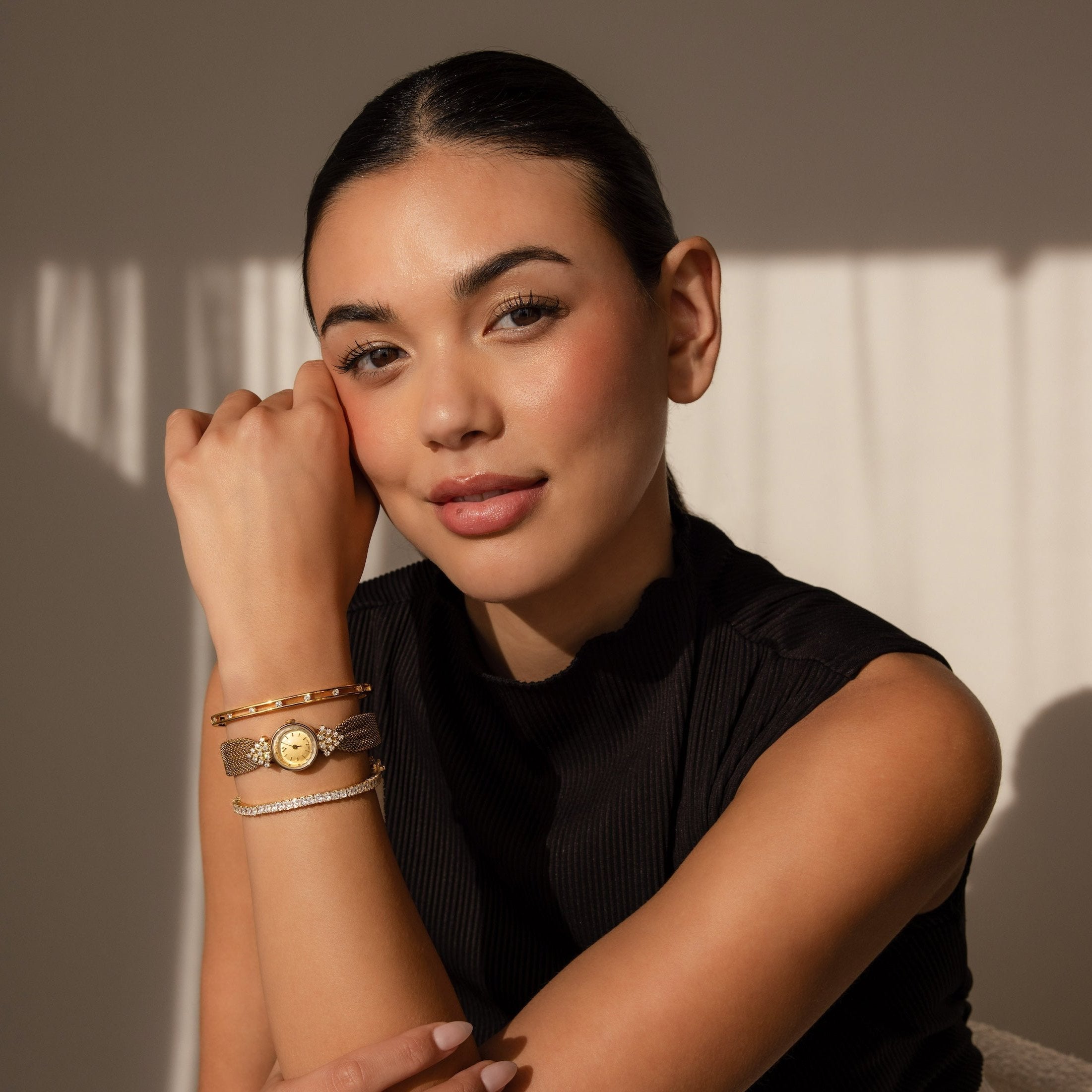 A woman in a black top poses with her hand near her face, highlighting layered gold and brown bracelets and the Diamond Mesh Timepiece under soft lighting.