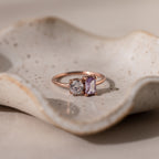 The Toi et Moi Emerald Birthstone Ring with a clear round gem and a purple rectangular gem is displayed on a textured ceramic dish.