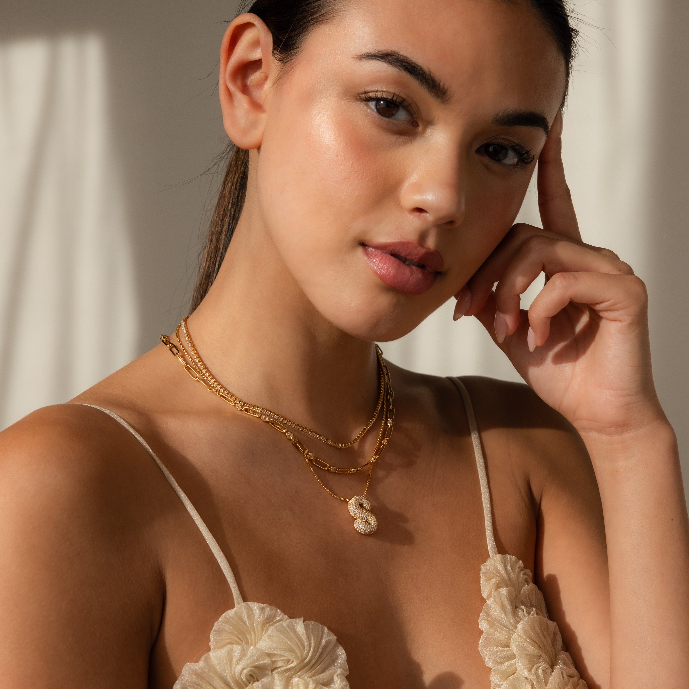 Woman wearing layered gold necklaces, including the Pave Puff Letter Necklace in 18K Gold, styled with a cream floral top and posing with her hand near her face.