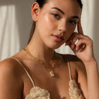 A woman wearing a Pave Puff Letter Necklace and a box chain, styled with a cream floral top, poses with her hand near her face.