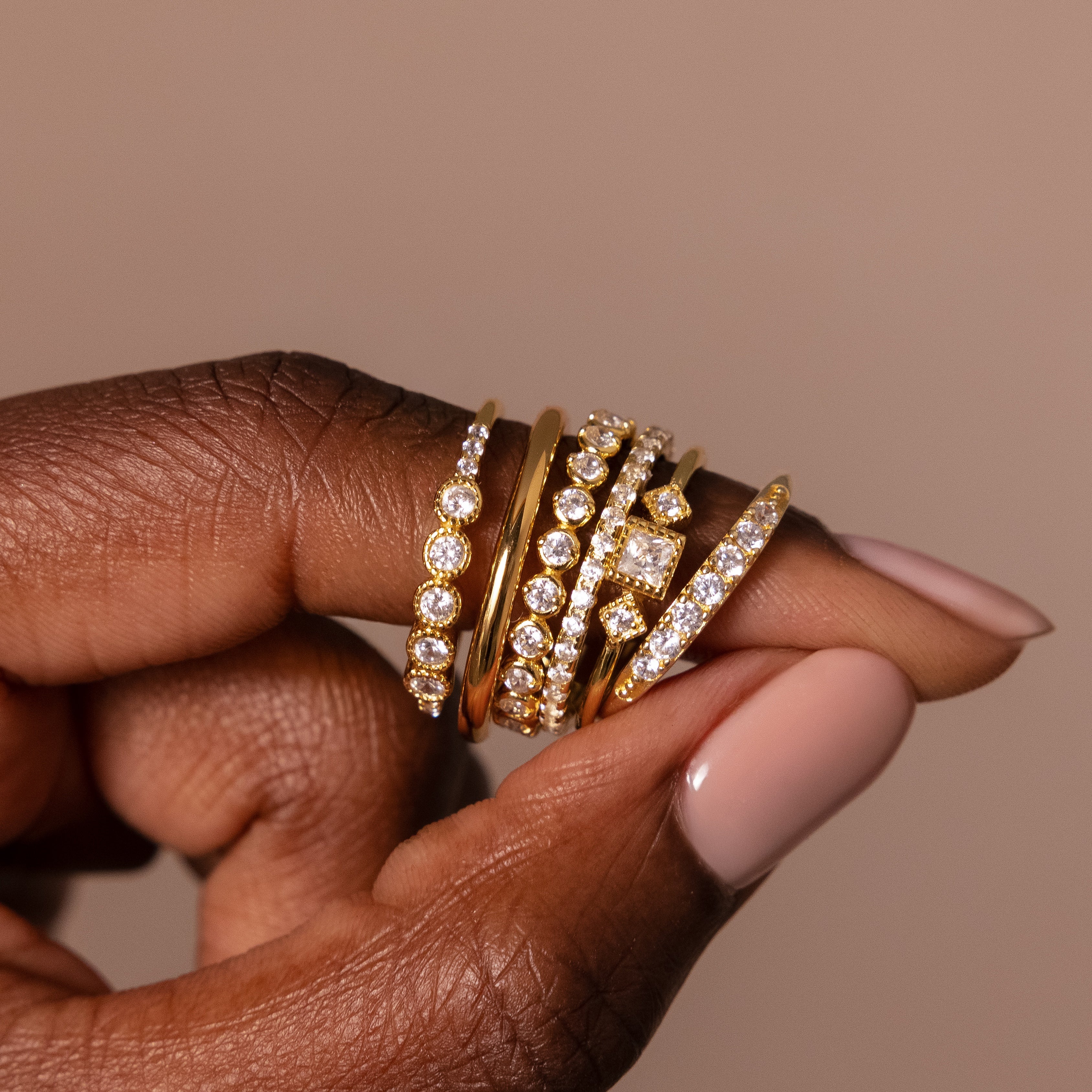 A hand with nude nails showcases the Art Deco Diamond Ring Set—five gold rings sparkling with diamonds—capturing vintage glamour against a neutral background.