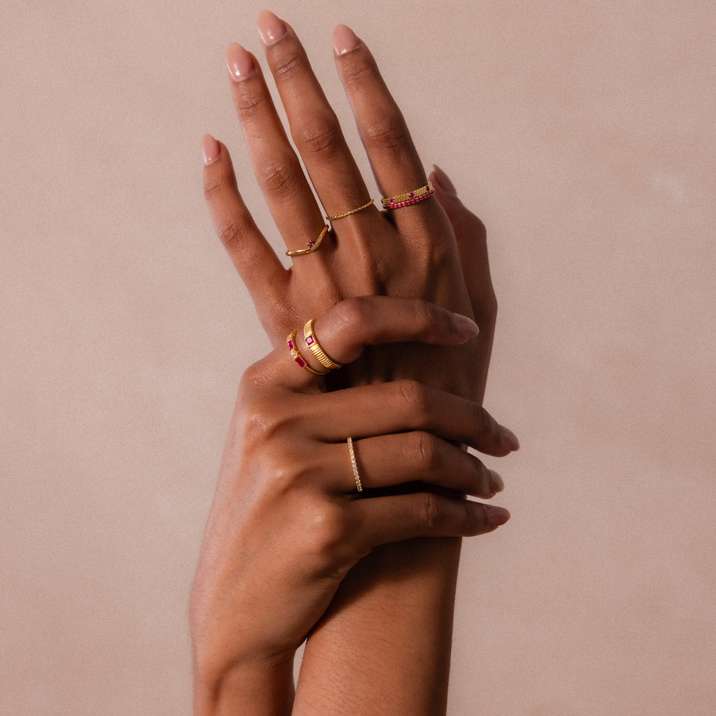 Two hands with manicured nails showcase multiple gold and gemstone rings, including a striking birthstone piece and the Ribbed Ruby Band, all against a beige background.