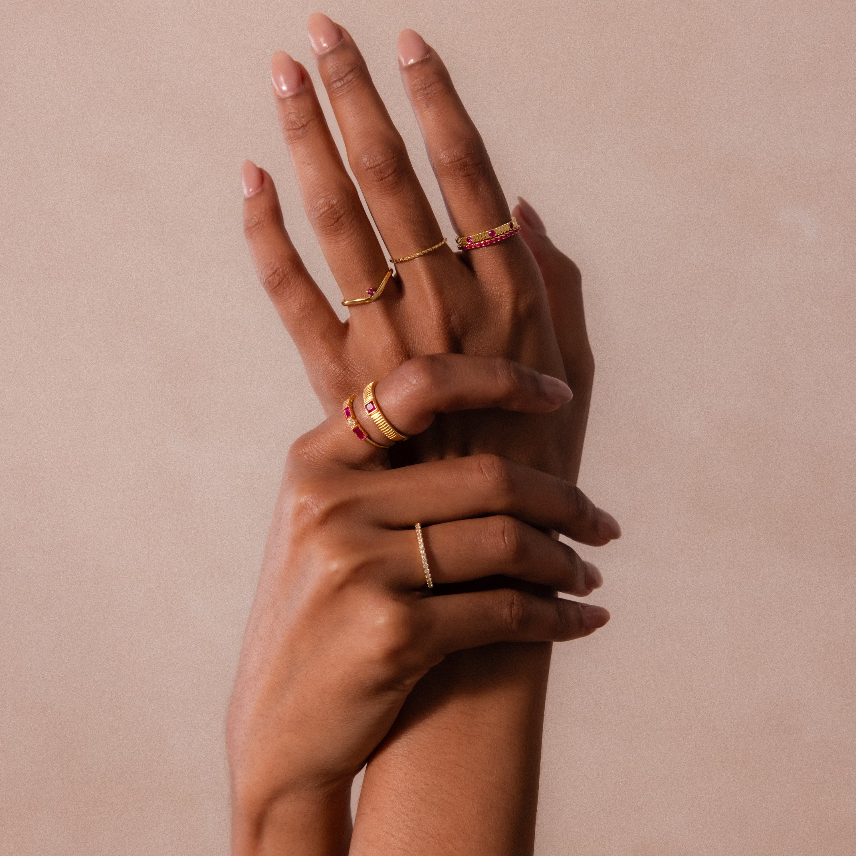 Two manicured hands wear multiple gold and red gemstone rings, including the Ribbed Topaz Band, against a beige background.