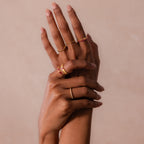 Two hands with manicured nails display several gold rings, including the Ribbed Blue Zircon Band, set against a soft beige background.