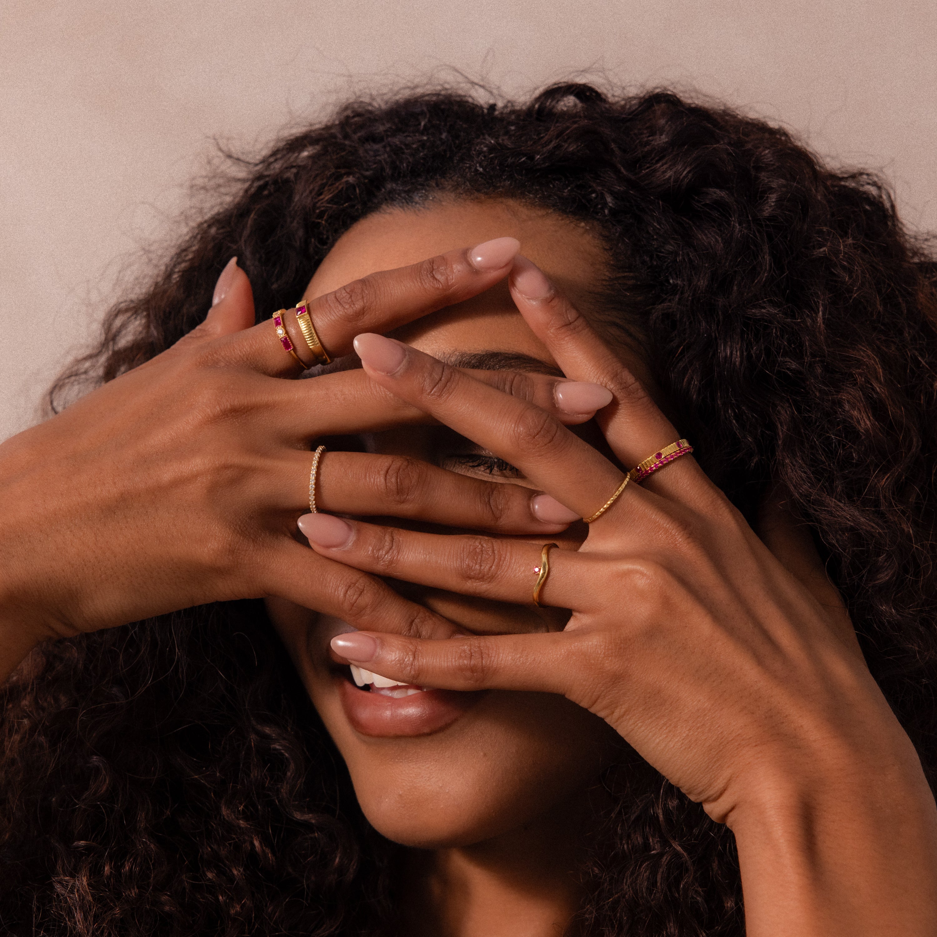 A woman with curly hair smiles, covering her face with hands adorned with Ribbed Topaz Band rings featuring gold ribbed bands and square-cut gemstone accents.