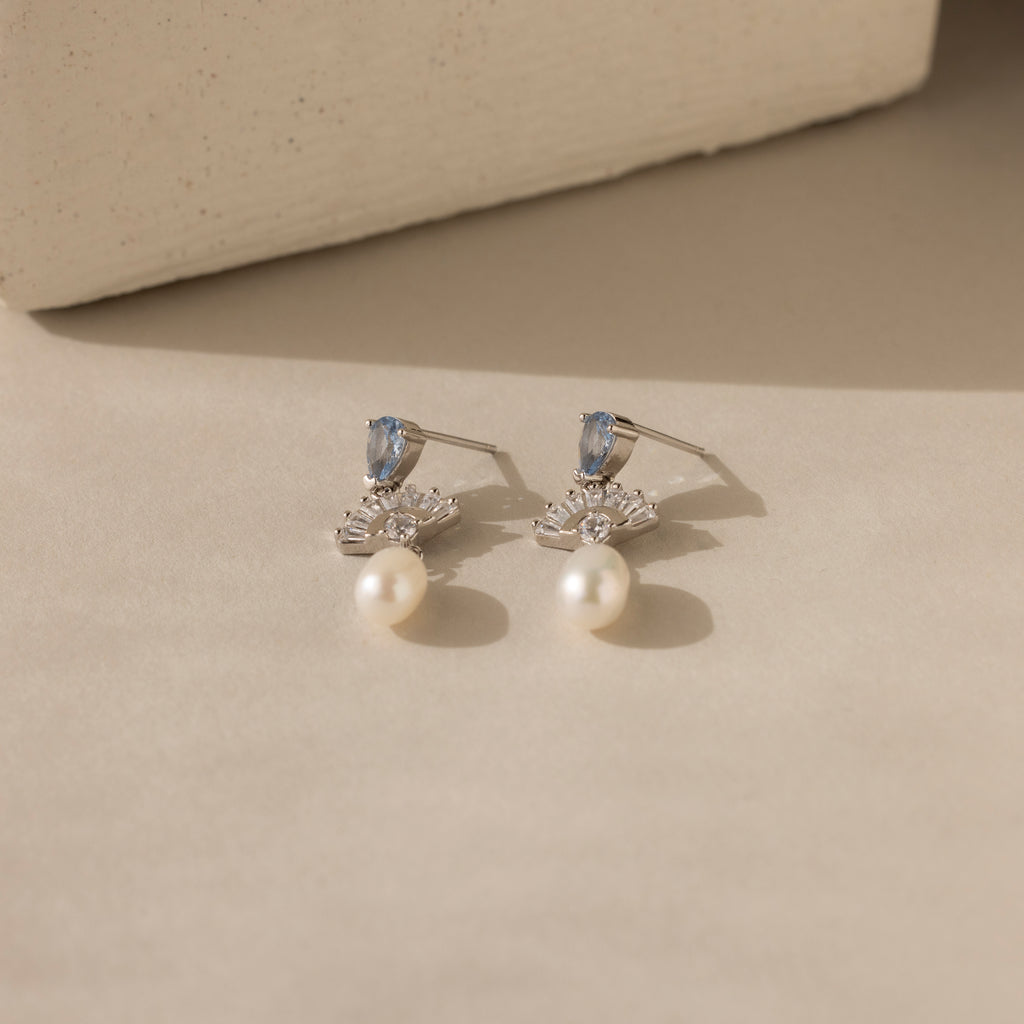 Aquamarine Pearl Drop Earrings in Silver, adorned with blue stones, clear crystals, and dangling pearls, exude vintage glamour on a beige surface with soft shadows.