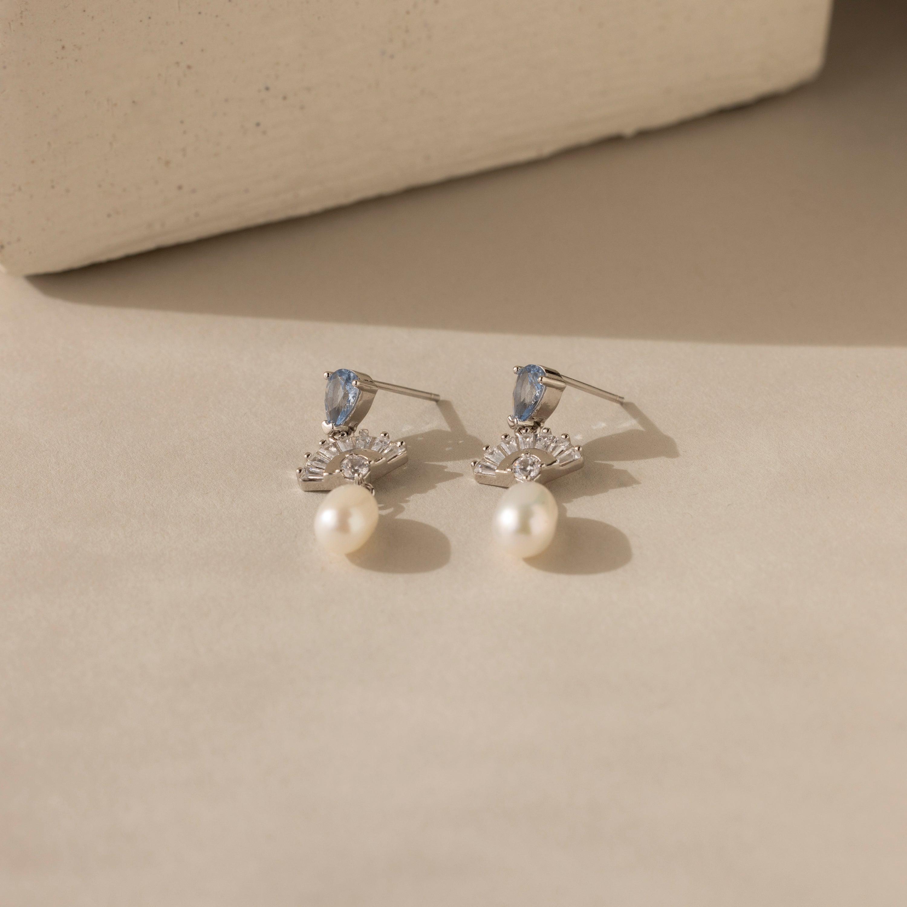 Aquamarine Pearl Drop Earrings in Silver, adorned with blue stones, clear crystals, and dangling pearls, exude vintage glamour on a beige surface with soft shadows.