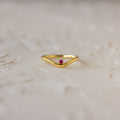 The Pink Ruby Drop Curve Ring, featuring a delicate red gemstone, is perfect as a birthstone ring and is displayed on a light textured surface.