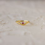 The Pink Ruby Drop Curve Ring, featuring a delicate red gemstone, is perfect as a birthstone ring and is displayed on a light textured surface.