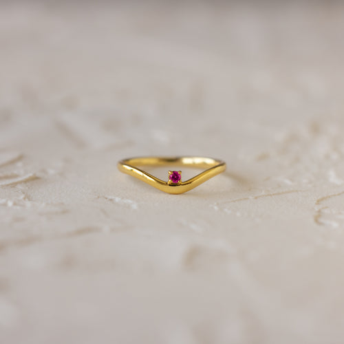 The Pink Ruby Drop Curve Ring, featuring a delicate red gemstone, is perfect as a birthstone ring and is displayed on a light textured surface.