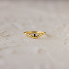 The Sapphire Drop Curve Ring, a gold stacking ring with a small dark sapphire gemstone, rests on a textured, light-colored surface.