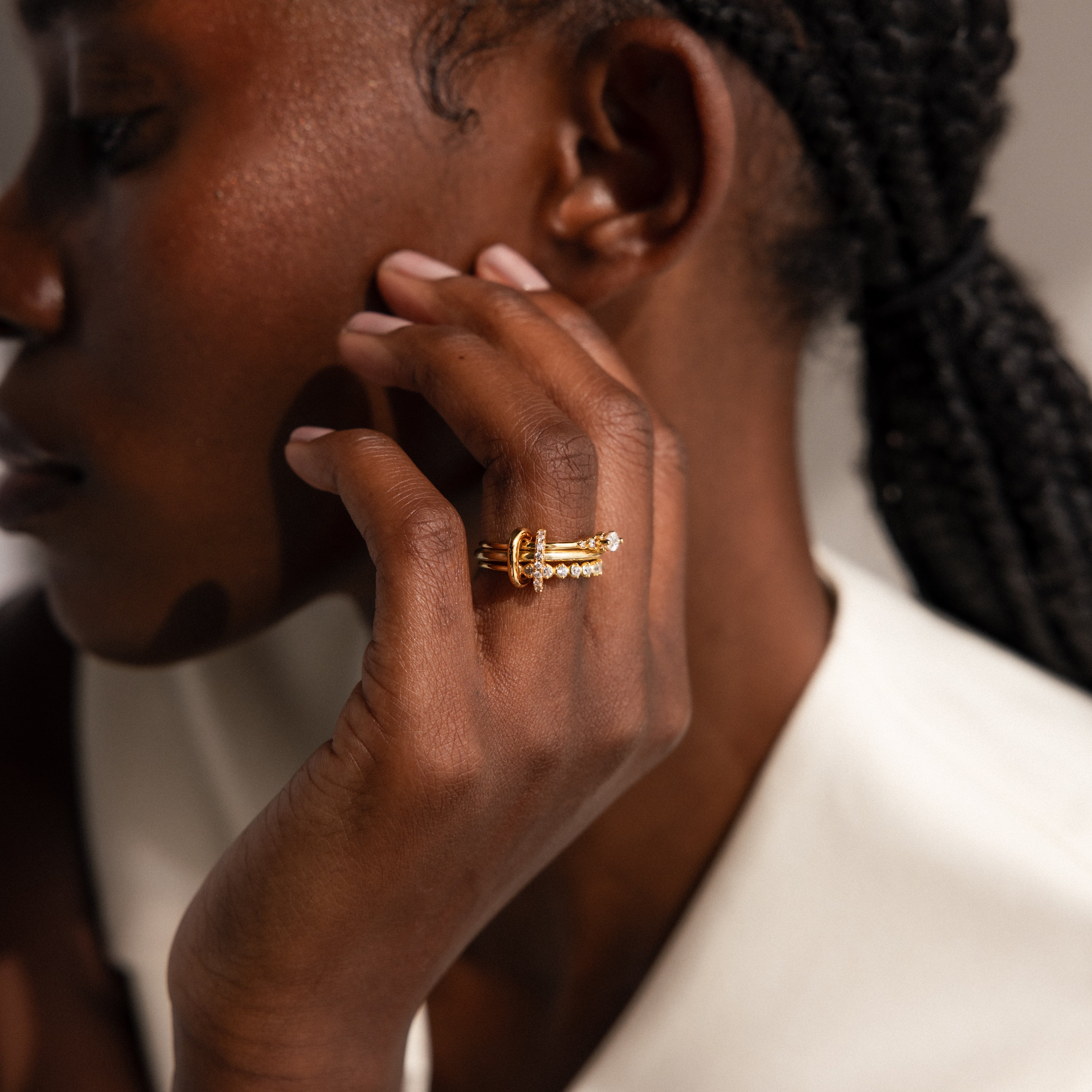 A person with braided hair wears a white top and touches their face, highlighting the Multipurpose Pave Ring Lock Set in 18K Gold—a customizable piece featuring a striking pavé-detailed clasp.