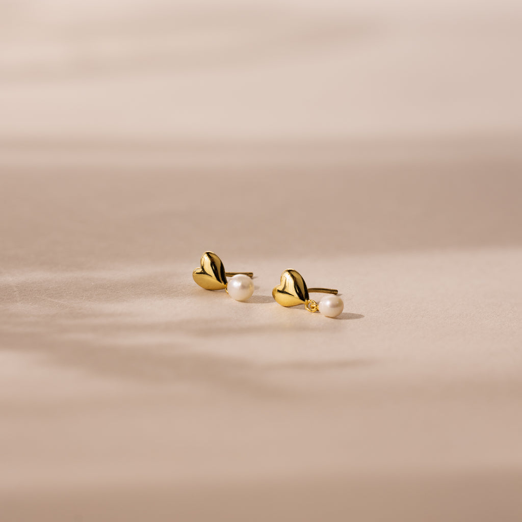 Heart Pearl Drop Earrings in 18K Gold, featuring delicate pearl accents, rest on a soft beige surface bathed in gentle sunlight.