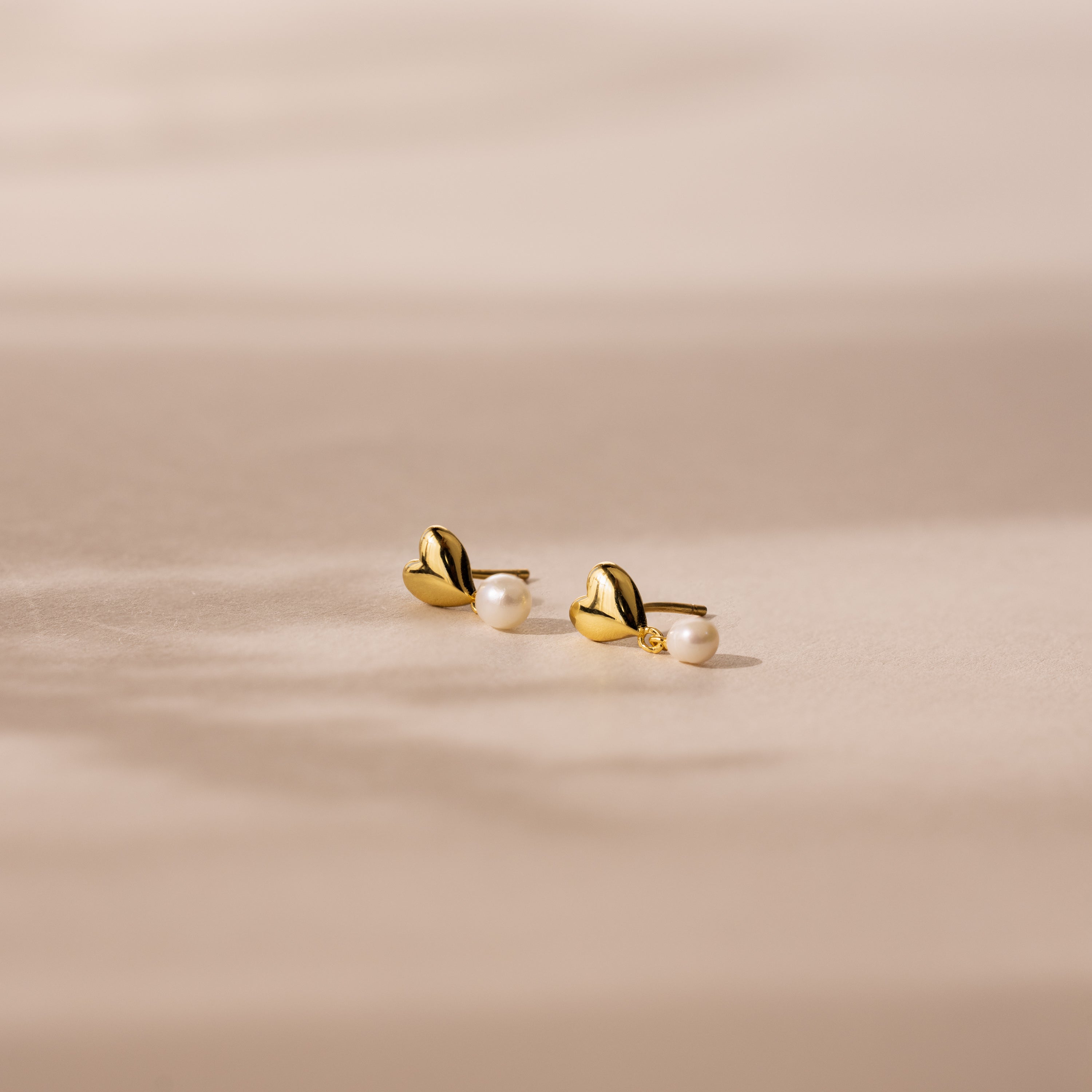 Heart Pearl Drop Earrings in 18K Gold, featuring delicate pearl accents, rest on a soft beige surface bathed in gentle sunlight.