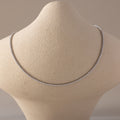 A Diamond Tennis Choker Necklace in white gold displayed on a beige textured neck form. The necklace features a continuous line of brilliant-cut white diamonds set in a sleek white gold setting, forming a minimalist and luxurious curve across the collarbone.