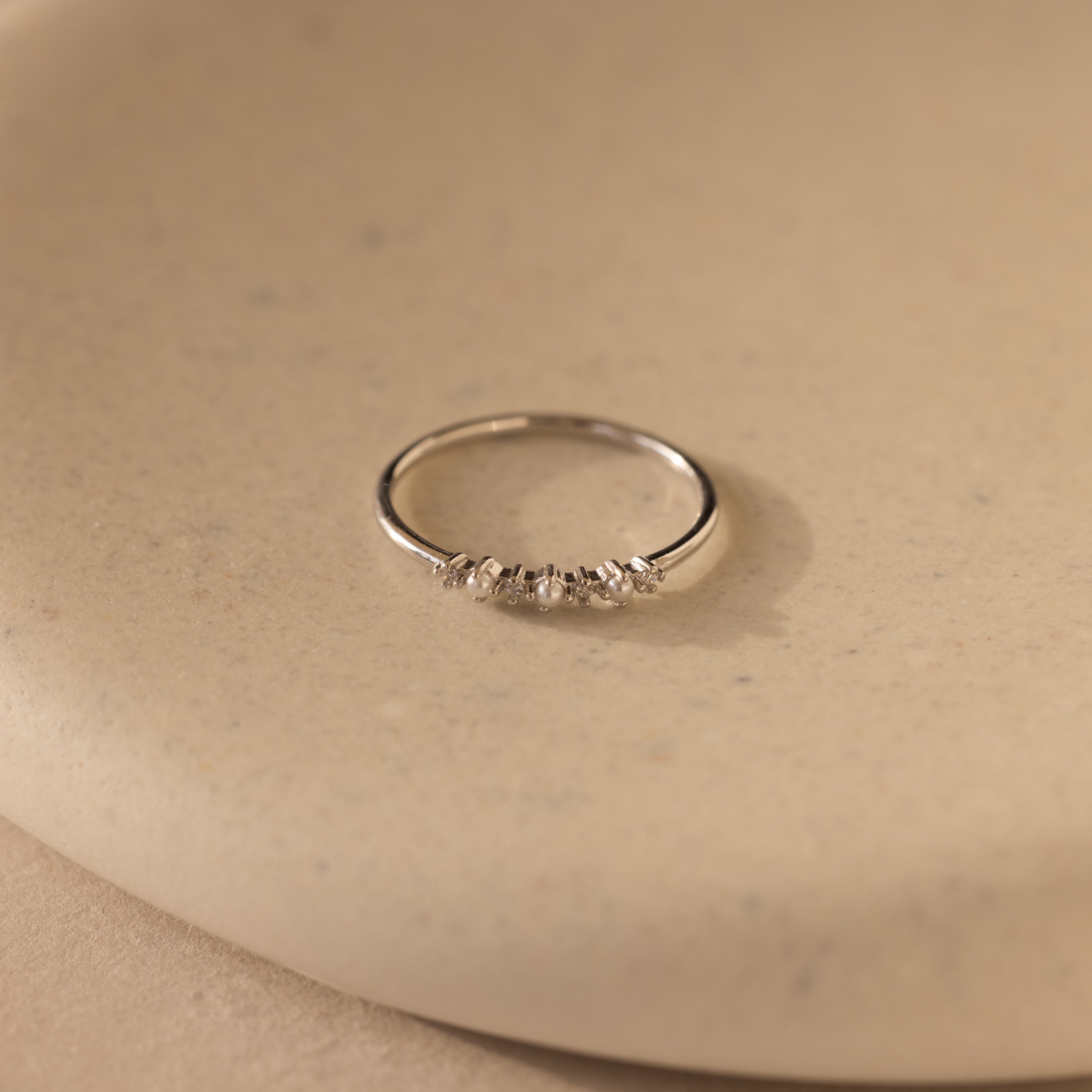 The Delicate Pearl Diamond Ring is a silver stacking ring featuring pearls and diamonds, displayed on a smooth beige surface.