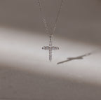 The Pave Cross Necklace in Silver features a sparkling pendant casting a shadow on a smooth surface—an exquisite piece of elegant jewelry.