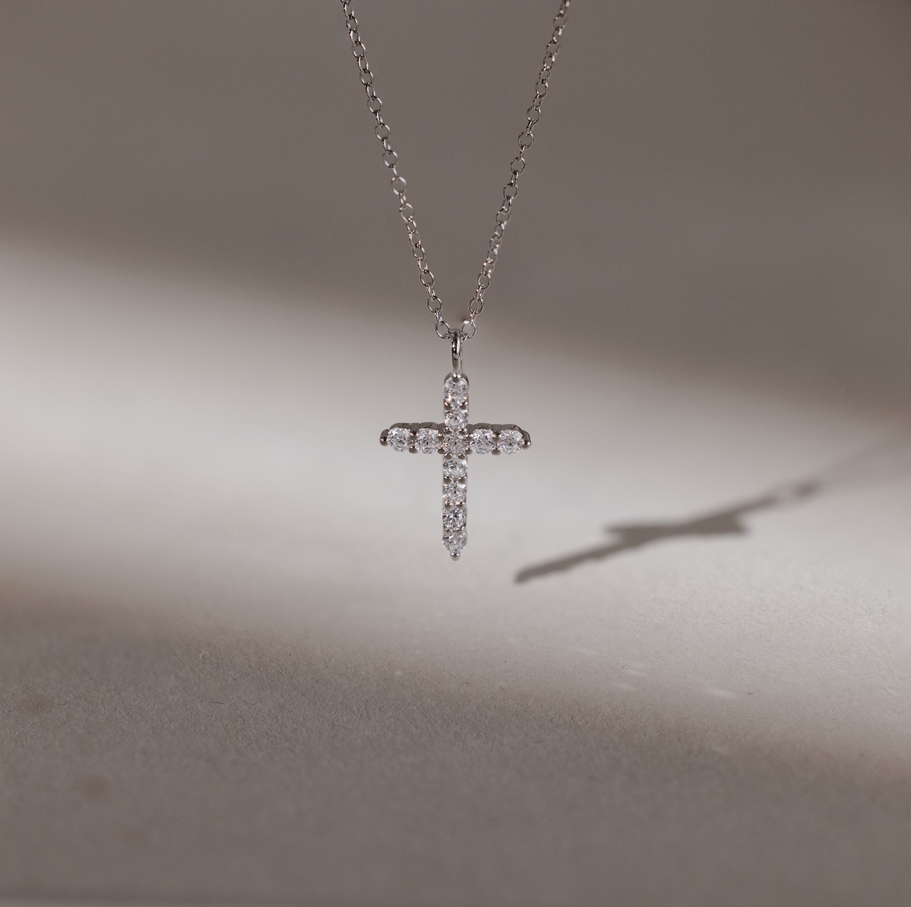 The Pave Cross Necklace in Silver features a sparkling pendant casting a shadow on a smooth surface—an exquisite piece of elegant jewelry.