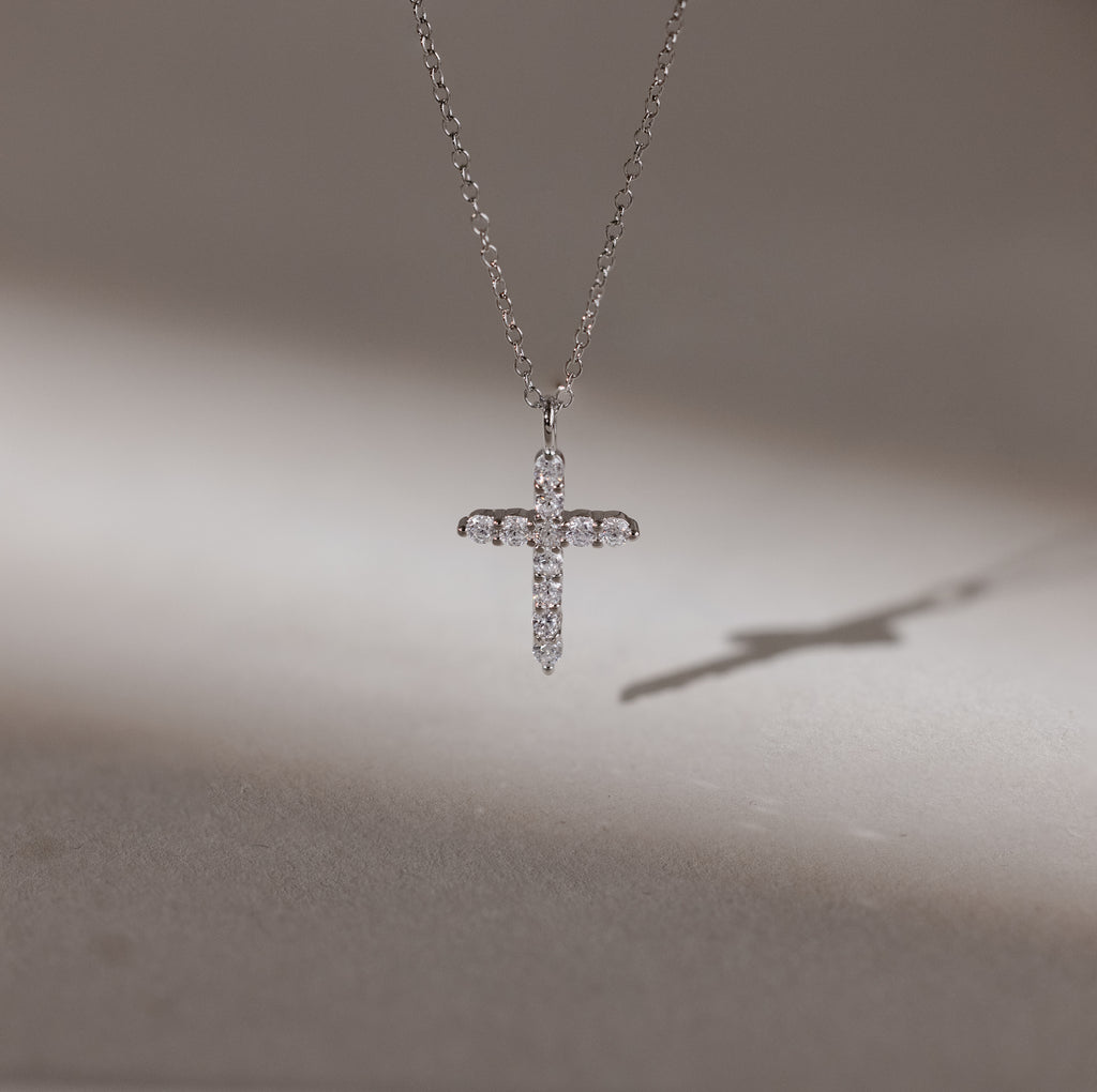 The Pave Cross Necklace shimmers with embedded diamonds, casting a delicate shadow on a smooth surface—an exquisite and elegant jewelry piece.