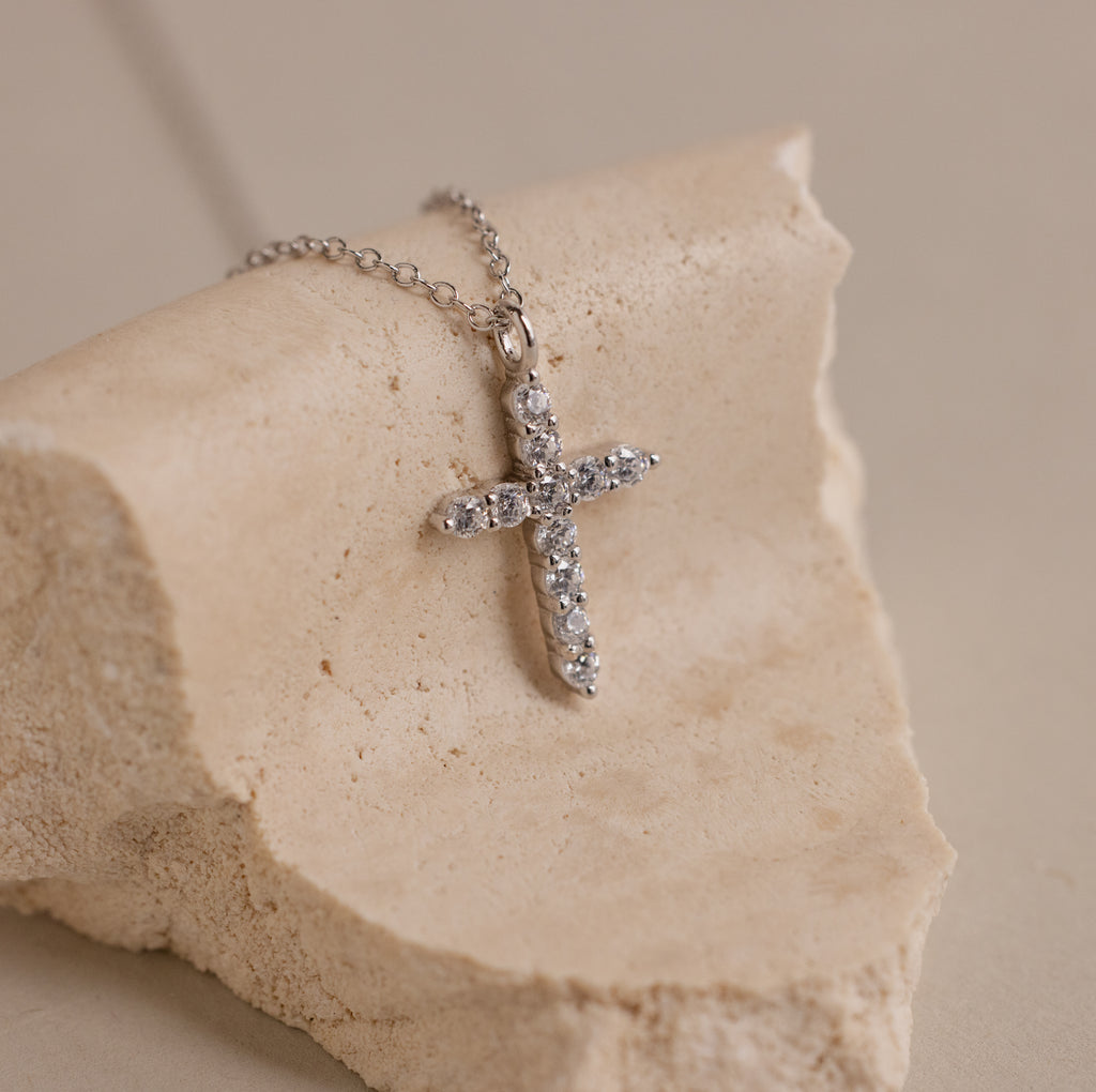 The Pave Cross Necklace in Silver features a diamond-accented cross and is elegantly displayed on a textured beige stone.