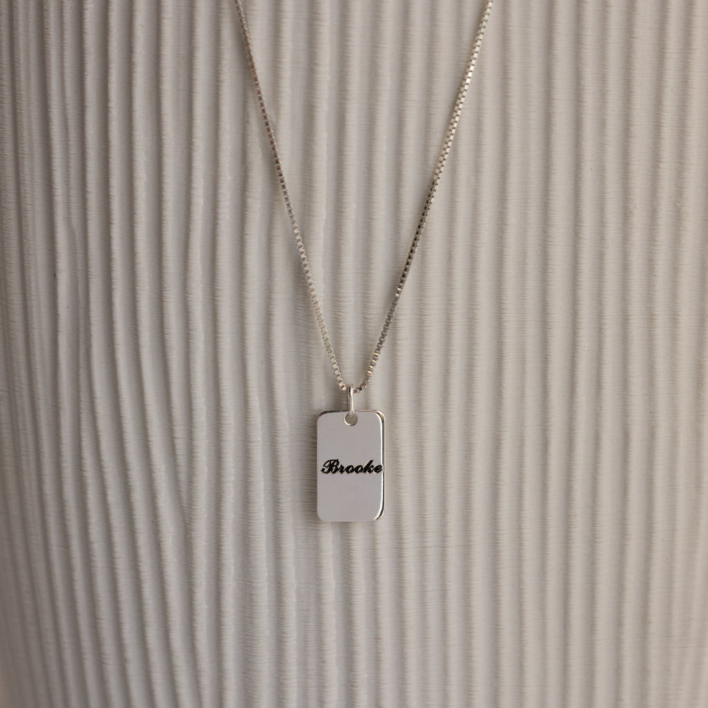 Name Tag Necklace in Sterling Silver with a rectangular pendant engraved "Brooke," shown on a textured white background.