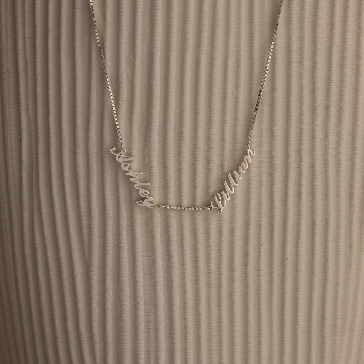 Necklace with 'Ashley' and 'Lillian' name charms in cursive on a textured brown background