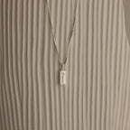 The Dainty Tag Name Necklace in Sterling Silver, featuring engraved text on a delicate chain, is displayed against a textured, light-colored background.