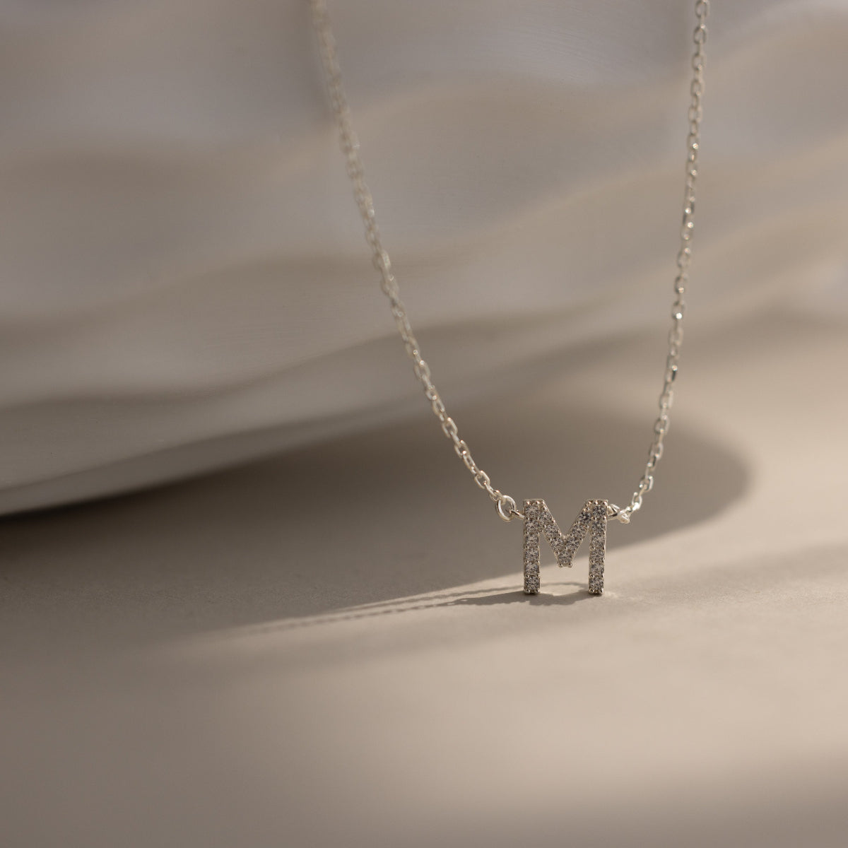 The Pave Initial Necklace features a sparkling, diamond-studded letter M pendant in silver, displayed on a light textured surface—a refined and personalized jewelry piece.