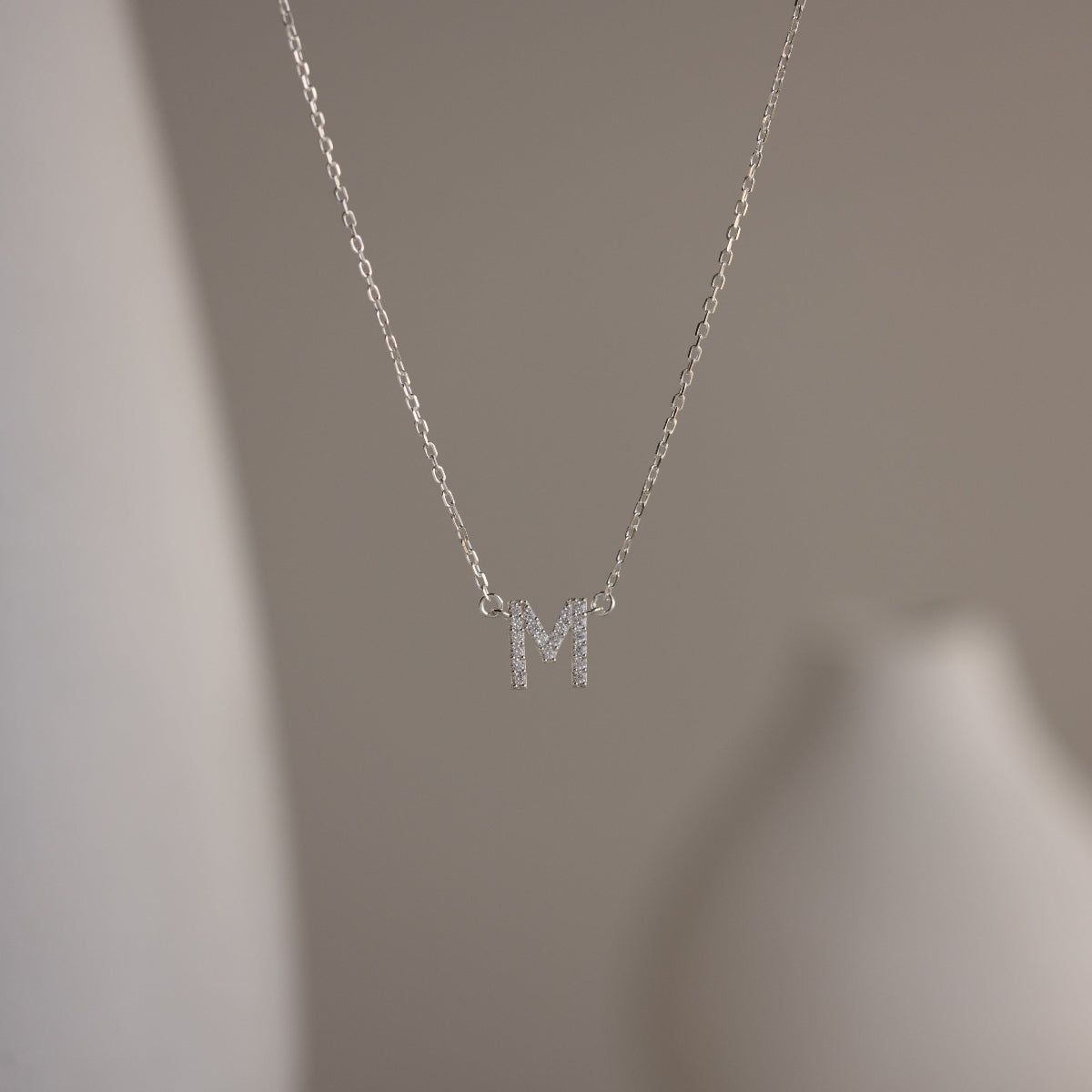 The Pave Initial Necklace features a sparkling letter M pendant, adding personalized elegance to your look against a blurred neutral background.