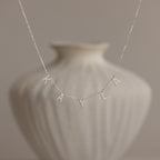 The Pave Letter Necklace in Sterling Silver, featuring charms that spell "KAYLA," is displayed against a blurred white vase, showcasing personalized elegance.