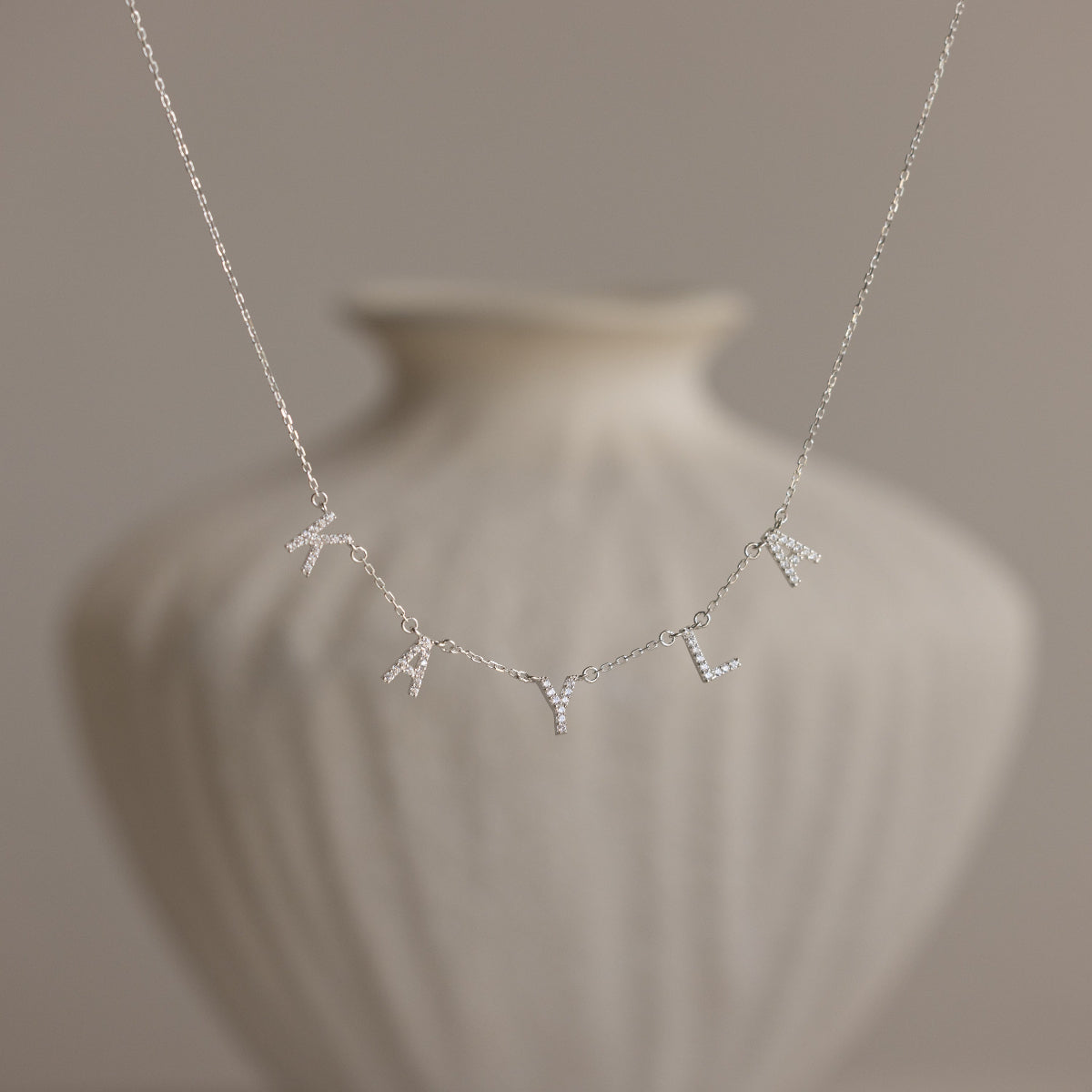 The Pave Letter Necklace in Sterling Silver, featuring charms that spell "KAYLA," is displayed against a blurred white vase, showcasing personalized elegance.