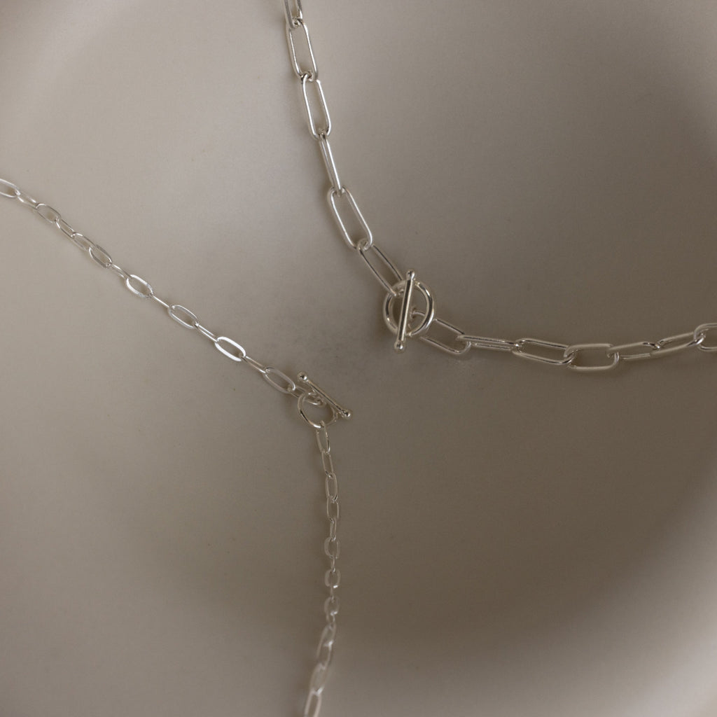 Three sterling silver chain necklaces, including a boho-chic layered piece and the Paperclip Toggle Necklace in Sterling Silver, are displayed on a light, curved surface.