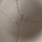 Three sterling silver chain necklaces, including a boho-chic layered piece and the Paperclip Toggle Necklace in Sterling Silver, are displayed on a light, curved surface.