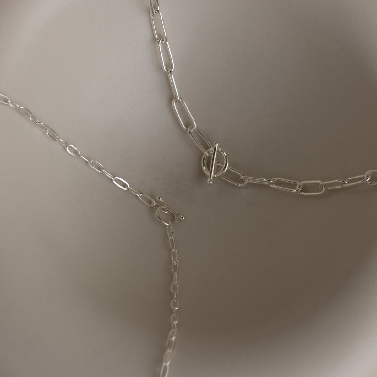 Three sterling silver chain necklaces, including a boho-chic layered piece and the Paperclip Toggle Necklace in Sterling Silver, are displayed on a light, curved surface.