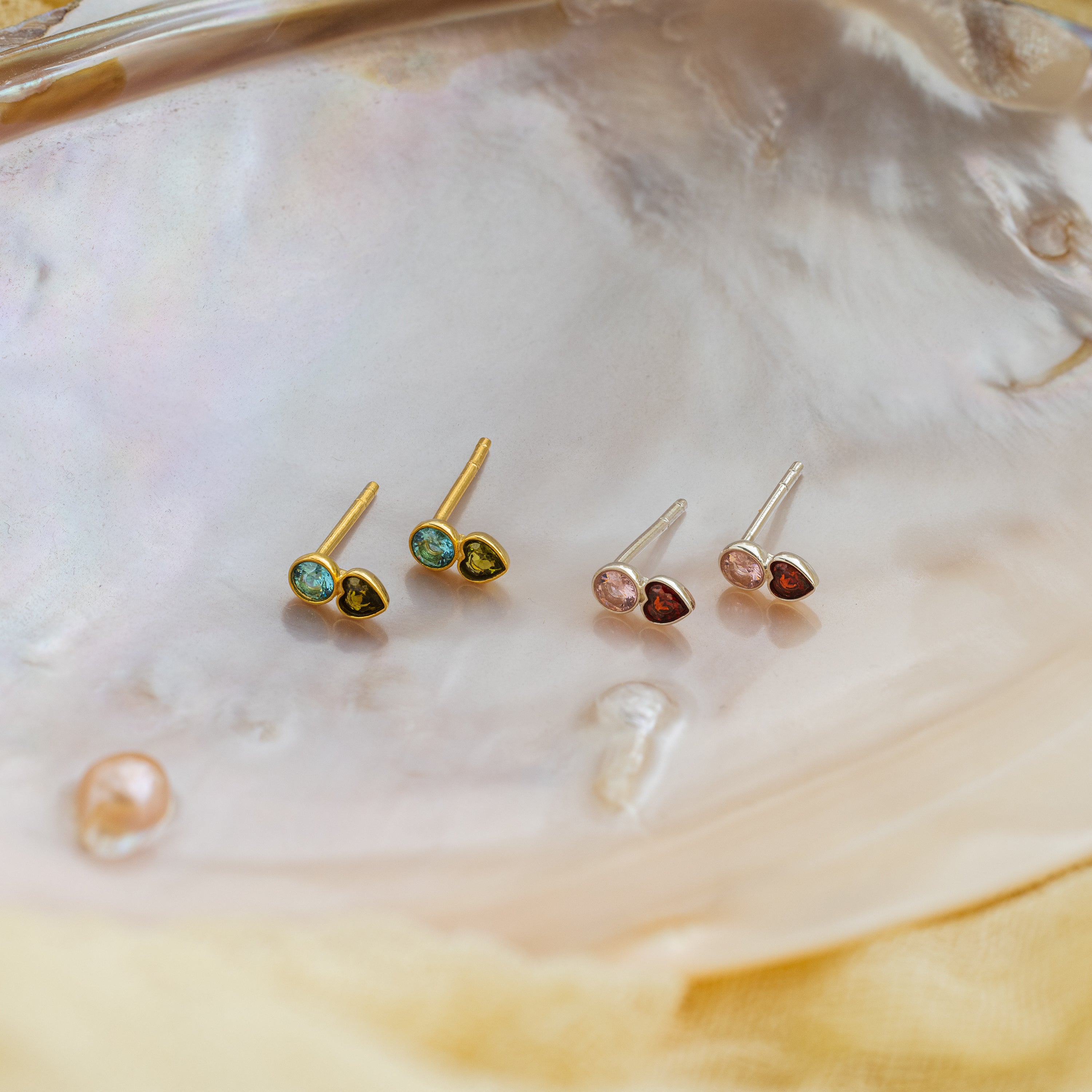 Two pairs of Toi et Moi Birthstone Stud earrings in gold and silver, displayed on a pearlescent shell surface—a perfect personalized gift for any occasion.