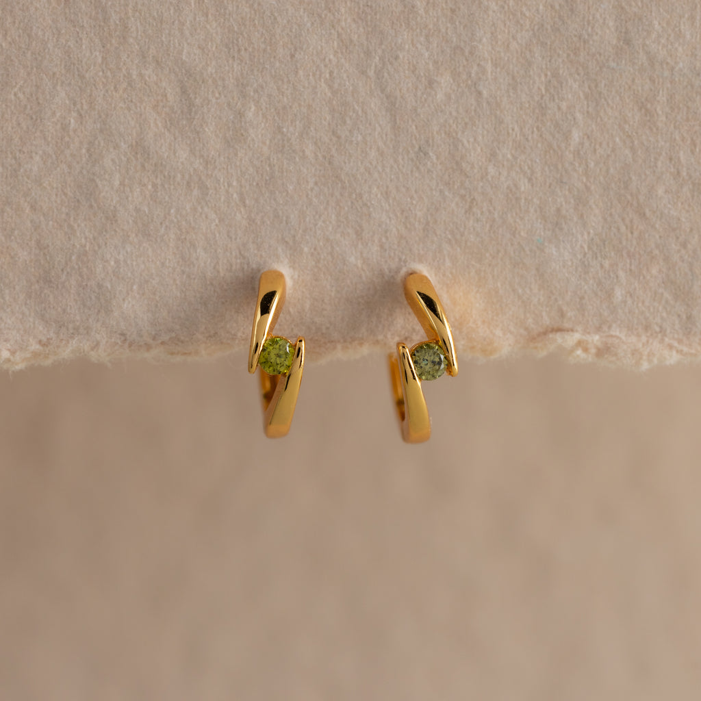 Small gold hoop earrings with green gemstones, shown on a beige surface—these Floating Peridot Huggies add a personalized jewelry touch to any look.