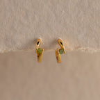 Small gold hoop earrings with green gemstones, shown on a beige surface—these Floating Peridot Huggies add a personalized jewelry touch to any look.
