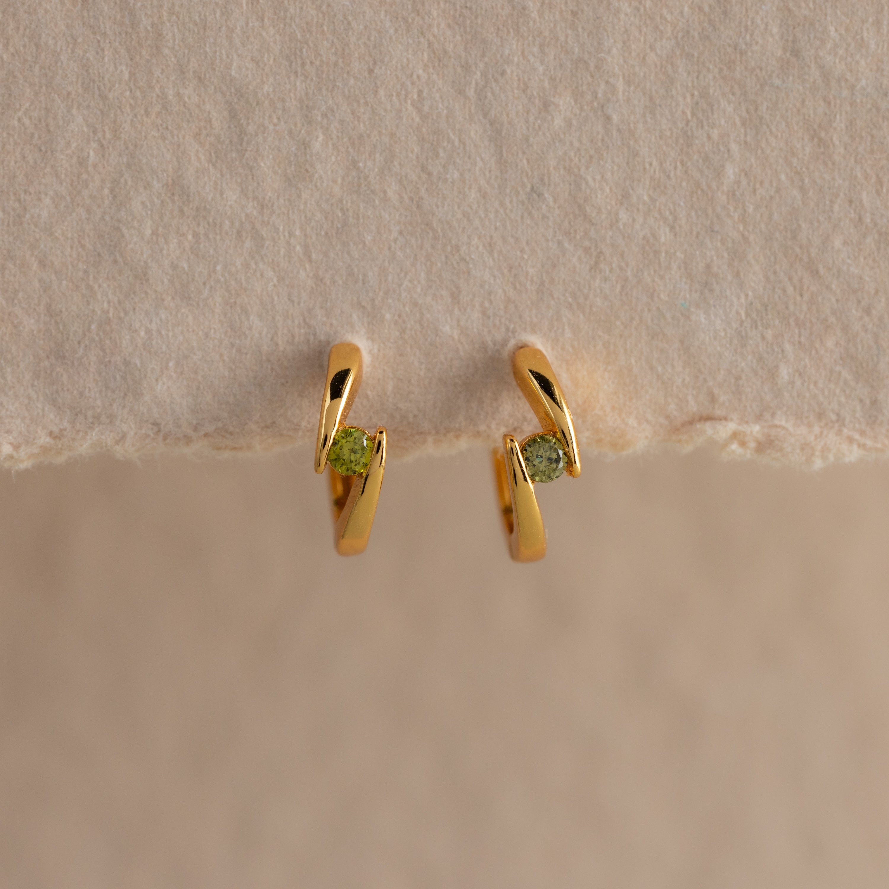 Small gold hoop earrings with green gemstones, shown on a beige surface—these Floating Peridot Huggies add a personalized jewelry touch to any look.