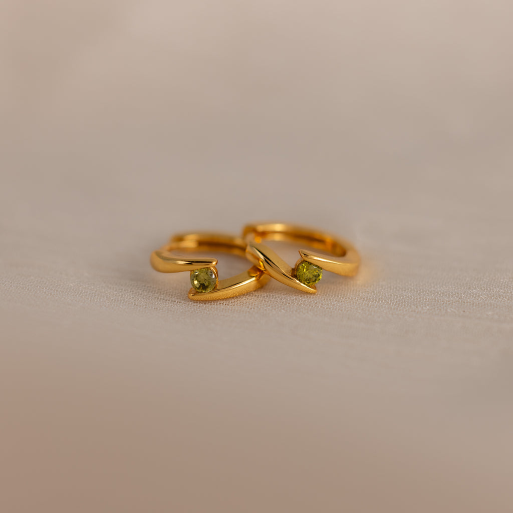 Two Floating Peridot Huggies, gold hoops with small green gemstones, are placed side by side on a light beige surface.